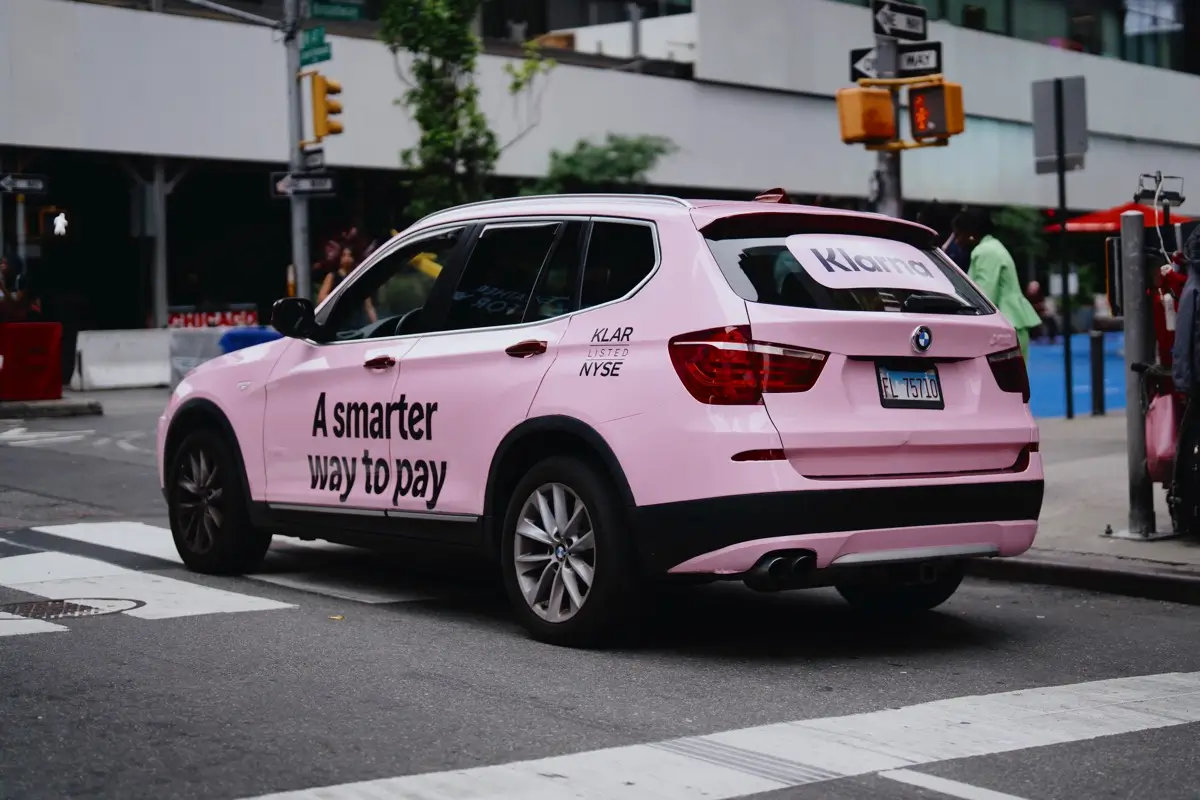 Pink SUV with "A smarter way to pay" and "Klarna" text on New York City street.